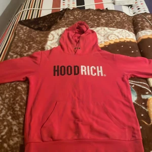 Röd hoodie från Hoodrich - Säljer en röd hoodie från Hoodrich med stor logga i svart och vitt på bröstet. Tröjan har huva med snörning och en stor magficka. Perfekt för dig som gillar streetwear och vill sticka ut med en färgstark look.
