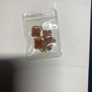 Guldarmband med röda fyrklöver - Stilrent armband med fem röda fyrklöverformade hängen infattade i guldfärgad metall. Kedjan är tunn och smidig med justerbar längd och ett diskret lås. Perfekt accessoar för att lyfta din outfit med en färgklick och trendig design.