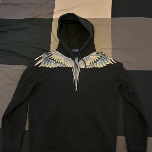 Svart hoodie med vingar från Marcelo Burlon - Svart hoodie från Marcelo Burlon County of Milan med ikoniskt vingtryck i grått och blått över axlar och bröst. Den är äkta, köpts från minto.se