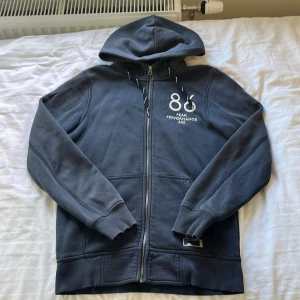 Mörkblå hoodie Peak Performance  - Mörkblå hoodie från Peak Performance med dragkedja och huva. Snyggt broderad logga och siffran 86 på bröstet. Mjukt fleecefoder på insidan och ribbade muddar. Perfekt för chill eller träning, med praktiska fickor och snörning i huvan.storlek S men sitter som M. Passar 170-178.