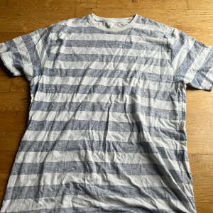 Randig t-shirt från Dressmann XL - Säljer en casual t-shirt från Dressmann i storlek XL. T-shirten har breda horisontella ränder i vitt och ljusgrått, klassisk rund hals och korta ärmar. Perfekt för en chill och avslappnad stil. Materialet är mjuk bomull som känns skönt mot huden.