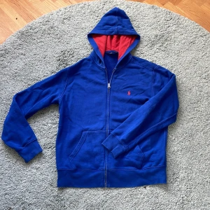 Blå hoodie från Polo Ralph Lauren - Säljer en blå hoodie från Polo Ralph Lauren med röd insida på huvan och rött broderat logotyp på bröstet. Tröjan har dragkedja framtill, känguruficka och är tillverkad i mjuk bomull. Perfekt för dig som gillar klassisk streetstyle. Kom gärna med frågor! 