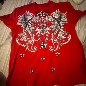Röd t-shirt med stjärntryck - Röd t-shirt med coolt tryck i svart, vitt och grått framtill. Designen har stora grafiska mönster och flera stjärnor på framsidan. Klassisk passform med rund hals och korta ärmar den var lång armad inann men jag klippte bort det. Perfekt för dig som vill sticka ut med en unik look. 