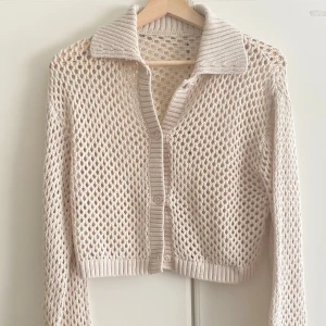 Beige stickad kofta med krage - Säljer en beige stickad kofta med hålmönster och ribbad krage. Koftan är croppad, har långa ärmar och knäpps framtill med knappar. Perfekt för lager-på-lager och ger en chill vibe till din outfit.