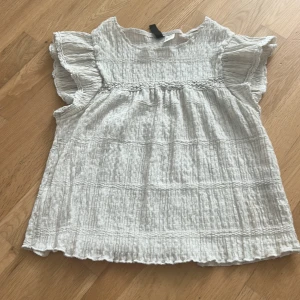 Vit blus med volangärm från Divided - Supersöt vit blus från Divided i storlek S. Blusen har en krinklad struktur, volangärmar och en lös passform. Perfekt för en fräsch och luftig look. Tillverkad i ett lätt material som känns skönt mot huden.