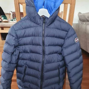 Mörkblå Colmar pufferjacka med huva - Säljer en mörkblå pufferjacka från Colmar med huva och blått foder. Jackan har dragkedja framtill, två fickor med dragkedja och en klassisk logga på ärmen. Supervarm och passar även till skidåkning. Använt ganska mycket, fortfarande inga skavanker.