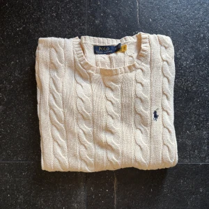 Kabelstickad beige tröja från Polo Ralph Lauren - Kabelstickad beige tröja från Polo Ralph Lauren med klassisk rund halsringning och broderad marinblå logga på bröstet. Tröjan är långärmad och har ribbade muddar och nederkant. Perfekt för lager-på-lager och snygg till jeans.