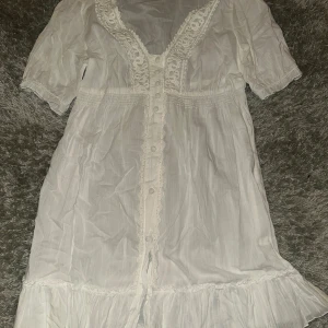 Vit kortklänning med spets från Vero Moda - Supersöt vit kortklänning från Vero Moda med spetsdetaljer längs v-ringningen och knappar framtill. Klänningen har korta ärmar, volangkant nedtill och är gjord i ett lätt bomullstyg som känns luftigt och fräscht.