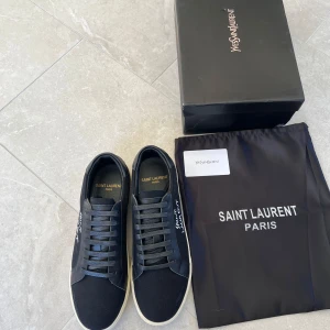 Svarta Saint Laurent sneakers - Snygga svarta sneakers från Saint Laurent med vit logga broderad på sidan. Skorna har ovandel i canvas och detaljer i skinn, samt vita gummisulor. Klassisk låg modell med snörning och rund tå. Perfekta för dig som gillar stilrena och exklusiva sneakers.