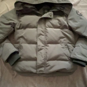 Canada Goose Jacka - Säljer en grå dunjacka från Canada Goose med huva och klassisk patch på ärmen. Jackan har dragkedja och knappar framtill, stora fickor och ribbade muddar vid ärmsluten. Perfekt för kalla dagar och riktigt snygg streetstil.