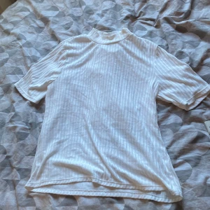 Vit ribbad T-shirt från H&M - Vit ribbad polotopp från H&M i storlek L. Toppen har korta ärmar och en högre krage, med ett diskret randigt mönster i tyget. Perfekt att styla med jeans eller kjol för en clean look.
