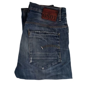  G-Star RAW GS01 Jeans - Snygga blå jeans från G-Star RAW, modell GS01. Jeansen har klassisk femficksdesign, coola slitningar och kontrastsömmar. Märkespatch i läder baktill och raka ben. Perfekta för dig som gillar streetwear och avslappnad stil.