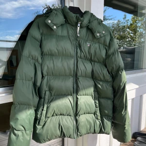 Grön pufferjacka från Gant - Snygg grön pufferjacka från Gant med huva och dragkedja framtill. Jackan har en quiltad look, två fickor med dragkedja och en liten logga på bröstet. Perfekt för kyliga dagar och riktigt skön att bära.