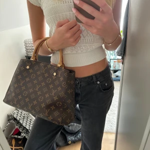 Handväska/Väska - Säljer en klassisk handväska i brunt monogrammönster med guldiga detaljer och röda innerfickor. Väskan har både handtag och axelrem, vilket gör den superflexibel. Tillverkad i skinn och canvas, perfekt för dig som vill ha en ikonisk och snygg accessoar. Aldrig använd.