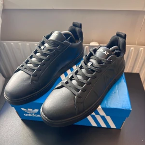 Svarta Replay sneakers med präglad logga - Snygga svarta sneakers från Replay med präglad REP och LAY på sidorna. Skorna har rund tå, platt sula och snörning. Tillverkade i skinnliknande syntetmaterial med svart textilfoder. Perfekt för dig som gillar stilrena och moderna detaljer. Limmat fram på ena tån men märks knappt