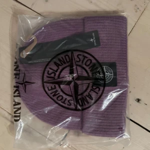 Lila mössa från Stone Island - Snygg lila mössa från Stone Island med ribbad struktur och klassisk logopatch framtill. Perfekt för dig som vill ha en cool och stilren accessoar till höst och vinter. Mössan är stickad och kommer i originalförpackning.