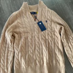 Beige stickad tröja Polo Ralph Lauren - Kabelstickad beige tröja från Polo Ralph Lauren med halv dragkedja och ribbad krage. Klassisk marinblå logga broderad på bröstet och snyggt läderdetalj på dragkedjan. Perfekt för lager-på-lager och chill höst- eller vinterstil.