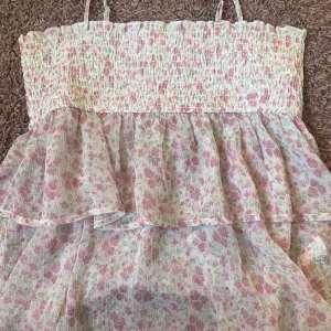 Blommig sommarblus Gina Tricot - Supersöt blommig klänning från Gina Tricot med smock upptill och tunna axelband. Klänningen är vit med rosa och gröna blommor, har volangdetaljer och är gjord i ett lätt, luftigt material. Perfekt för varma dagar och somriga vibes! Strl 158/164 men skulle säga att det funkar som XS
