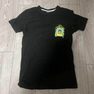 Svart Avenida Casablanca t-shirt - Svart t-shirt från Avenida Casablanca med färgglad tryckt grafik fram och bak. Stort motiv av ett fönster med utsikt över en byggnad och gröna blad, samt texten 'Avenida Casablanca'. Klassisk passform och rund hals. Perfekt statement-plagg.