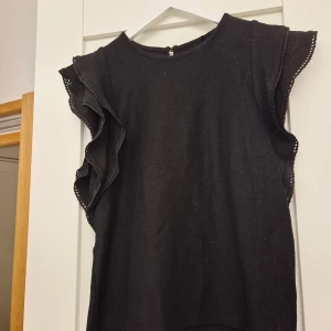 Svart volangblus från Holly & Whyte - Snygg svart blus från Holly & Whyte med volangdetaljer längs ärmhålen och dekorativ kant. Blusen är ärmlös och har en rund halsringning samt knapp i nacken. Perfekt för dig som gillar stilrena plagg med en twist.