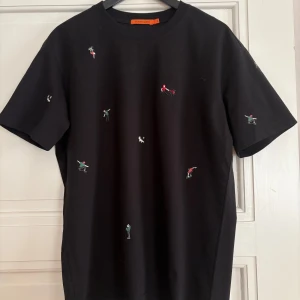 Svart t-shirt med broderier Gianni Lupo - Svart t-shirt från Gianni Lupo i bomull med små broderade figurer och hundar över hela plagget. Klassisk passform med rund hals och korta ärmar. Perfekt för dig som vill ha en unik och lekfull stil.