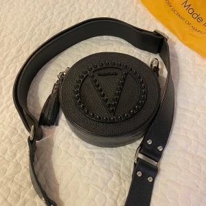Snygg rund axelväska från Valentino - Cool svart rund axelväska från Valentino med bred justerbar axelrem och snygga nitar i ett V-mönster på framsidan. Väskan är i äkta präglat skinn och har dragkedja med metallring där det står Valentino. Kommer med original dustbag och tags.