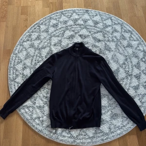 Svart kofta i ull från Suitsupply - Stilren svart kofta från Suitsupply i 100% ull. Modellen har hel dragkedja framtill, hög krage och långa ärmar. Perfekt för lager-på-lager och funkar året runt. Enkel och klassisk design som passar till det mesta.