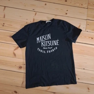 Svart Maison Kitsuné t-shirt - Svart t-shirt från Maison Kitsuné med vit tryckt text på bröstet: 'Maison Kitsuné Palais Royal Paris, France'. Klassisk passform, rund hals och korta ärmar. Storlek M (står L me passar som M). Axelbredd:45cm, längd:70cm. Tillverkad i 100% bomull för en skön och mjuk känsla. Inga defekter utom att taggen ser lite urtvättad ut.