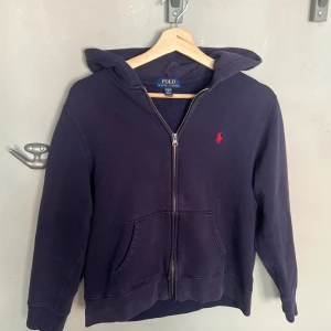 Mörkblå zip  hoodie från Polo Ralph Lauren - Mörkblå zip hoodie från Polo Ralph Lauren med dragkedja och huva. Klassisk röd broderad logga på bröstet och två stora fickor framtill. Tillverkad i mjuk bomullsmix, perfekt för chill dagar. Snygg och enkel att matcha med det mesta. Tröjan är storlek 14-16 så alltså ungefär xs i vuxen storlekar. Hör av er vid frågor eller så:)