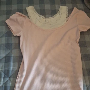 Ljusrosa ribbad topp från Lindex - Supersöt ljusrosa ribbad topp från Lindex med kort ärm och offshoulder-design. Under har den en vit spetskantad insydd del som ger lager-på-lager känsla. Perfekt för dig som gillar romantiska och feminina detaljer.