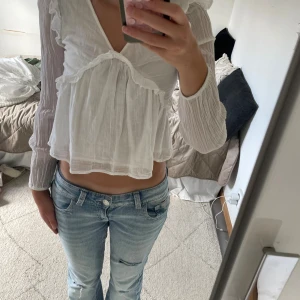 Vit volangblus från Gina Tricot - Supersöt vit blus från Gina Tricot med v-ringning, långa ärmar och volangdetaljer framtill och på axlarna. Blusen är lätt och luftig i tunt bomullstyg och har en lite croppad passform. Säljer på grund av att den tyvärr är lite för liten för mig