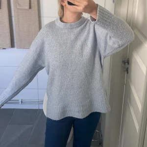 Ljusgrå stickad tröja med rund hals - Mysig ljusgrå stickad tröja med rund halsringning och avslappnad passform. Tröjan har långa ärmar och ribbade detaljer vid hals, ärmslut och nederkant. Perfekt för kyliga dagar och enkel att matcha med jeans eller kjol.