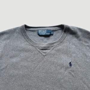 Ralph Lauren - Stickad Tröja - Storlek S - VARAN KAN KÖPAS DIREKT PÅ VÅR HEMSIDA, ettmarke.se MÄRKE: Ralph Lauren. FÄRG: Grå med mörkblå logga. STORLEK: S. AXELBREDD (SÖM TILL SÖM RAKT ÖVER): 50 cm. TOTALLÄNGD (FRÅN KRAGEN): 62 cm. BREDD (PLATT MÅTT): 52 cm. ÄRMLÄNGD 