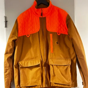 Carhartt-jacka, jakt/fiske/street - Nästintill oanvänd jacka från carhartt. Signalfärg på axlar och även på nedfällbar yta på ryggen. Rymliga fickor med plast invändigt för småvilt/fisk. 