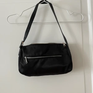 Väska  - Svart shoulder-väska från H&M. Stängs med dragkedja, ett fack på utsidan. Bredd: 16cm. Längd: 28cm. Reglerbart band. Defekt: missfärgat på knäppet vid bandet. Använd i okej skick annars. Bara att fråga om det är något du undrar! 