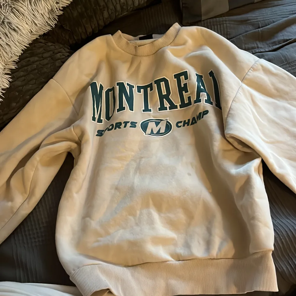 En beige sweatshirt från Gina Tricot med texten 'Montreal Sports Champ' i grönt på framsidan. Tröjan har långa ärmar och ribbstickade muddar va va vid ärmslut, nederkant och halsringning. De är en liten fläck på framsidan.. Tröjor & Koftor.