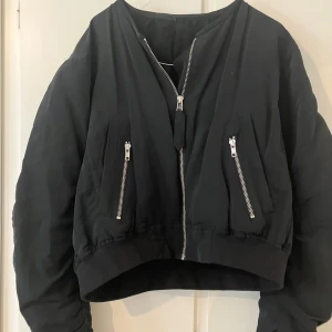 Svart bomberjacka från Acne Jeans - Snygg, svart bomberjacka från Acne Jeans med dragkedja framtill och två dragkedjeförsedda fickor på bröstet. Jackan har en kortare passform och är tillverkad i ett slitstarkt material.