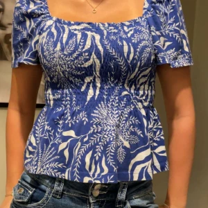 Blå blus med vitt bladmönster - En blå blus med vitt bladmönster. Blusen har korta ärmar och en fyrkantig urringning. Den är åtsittande upptill och har en lätt utställd nederdel.