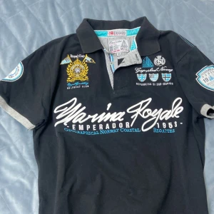 Svart pikétröja från Geographical Norway - En svart pikétröja från Geographical Norway med broderade detaljer och text på både fram- och baksidan. Tröjan har korta ärmar med gråa kanter och en krage med turkos insida. 