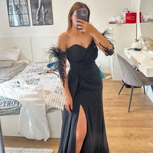 Klänning  - En elegant svart klänning med en slits i sidan. Klänningen har vackra fjädrar på axlarna och är perfekt för en festlig tillställning. Använt endast en gång. Köpt för 5000kr. Klänningen är i 36.