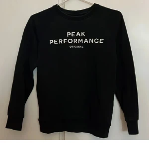 Svart tröja från Peak Performance - En svart tröja från Peak Performance med vit text 'PEAK PERFORMANCE ORIGINAL' på framsidan. Tröjan har långa ärmar och ribbstickade muddar vid ärmslut och nederkant. Passar bra för barn 
