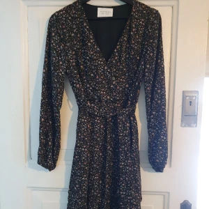 Klänning  - V-ringad,  volang nertill och resårband i ärmslut.  Dragkedja i sidan och band till midjan.