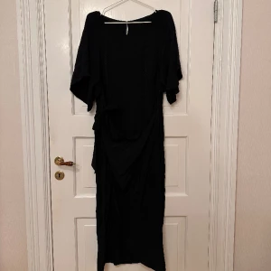 Svart klänning - Svart klänning med svarta prickar (se bild 2). Går ner till vaden. ”Omlott” i midjan då man knyter den där. Dragkedja i ryggen. Aldrig använd. 