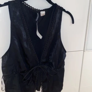 Västliknande topp - Väst inspirerad topp från H&M, aldrig använd (prislapp kvar). Glansigt material med knyte baktill för mer figurnära passform. Stl S