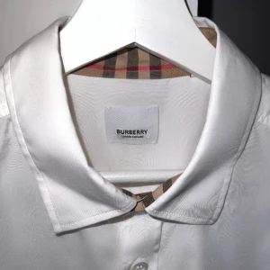 Burberry skjorta  - Storlek L, använd en gång. Tillverkad av 100% bomull, har denna skjorta en optisk vit rutig tryck, en klassisk krage och en knappknäppning framtill. De långa ärmarna med knappade manschetter och den böjda fållen ger en touch av sofistikering till den