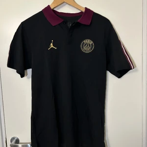 Jordan X PSG piké  - Jordan X Paris Saint-Germain piké med ränder vid ärmarna. I vinröd, svart och vit. Storlek S