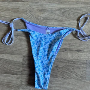 Blommig bikiniunderdel - En blommig bikiniunderdel från Bik Bok i storlek XL. Den har ett mönster av blå blommor på en ljusblå bakgrund och knytband på sidorna. Insidan är lila. Helt nya. 