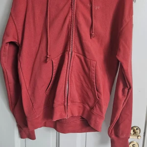 Röd hoodie från Levi's - En bekväm röd hoodie från Levi's med dragkedja och justerbar huva. Tröjan har långa ärmar och ribbstickade muddar vid ärmslut och nederkant. Den har också en känguruficka framtill.