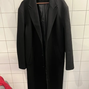 Svart trenchcoat  - En svart trenchcoat från Asos i storlek L. Köpt förra året. Produkten är knappt använd och i fint skick. Vill dock påpeka att denna kappa är väldigt lång. Jag är 178 cm och den går  nedanför knäna!