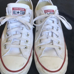 Vita Converse All Star sneakers - Ett par vita Converse All Star sneakers med snörning och röd rand runt sulan. Skorna har en klassisk design med Converse-logotypen på plösen och hälen.